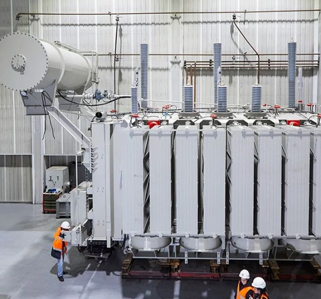 Three workers in safety vests and helmets examine a power transformer indoors.